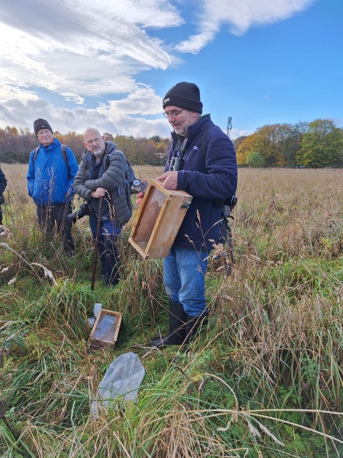 Person in a field holding a small mammal camera trap box to demonstrate a rmammal recording technique to a group of volunteers. Box is made of wood with a clear perspex lid.
